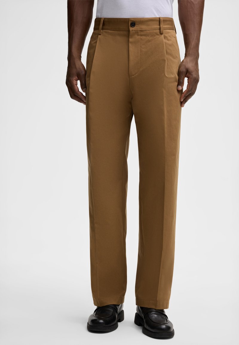 Pantaloni di cotone marroni con un design a gamba dritta, dotati di un unico bottone di chiusura e tasche laterali, indossati con scarpe nere.