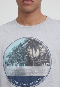Camiseta gris de manga corta con un gráfico circular de palmeras en azul y negro, superpuesto con rayas blancas y el texto "REIVINDICA TUS DERECHOS".