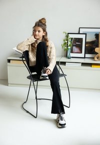 Beige kabelgebreide trui, zwarte flared broek en zwart-witte sneakers. Zittend op een zwarte stoel, met een lichte achtergrond en decor.