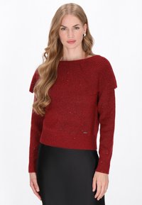 Pull en tricot rouge avec une texture côtelée, décolleté bateau et design superposé aux épaules, présentant un léger éclat sur l'ensemble.