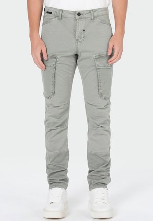 Homme portant un pantalon cargo slim gris clair avec poches avant à rabat et des baskets blanches, debout devant un fond uni.