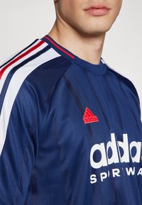 Granatowa koszulka sportowa z czerwonymi i białymi paskami na ramionach, z logo adidas w bieli i małym czerwonym emblematem na piersi. Gładka tekstura.