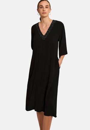 Femme portant une robe midi noire à manches mi-longues et un décolleté en V scintillant, debout avec une main dans la poche de la robe.