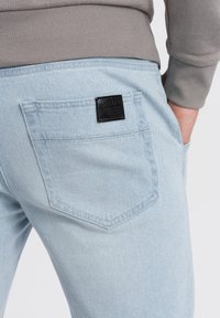 Jasnoniebieskie dżinsy z tylną kieszenią, zdobioną czarną skórzaną łatką. Tkanina jest gładka i lekko teksturowana, z subtelnym przeszyciem.