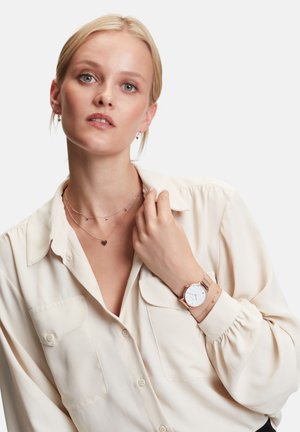 Roségoldene Uhr mit weißem Zifferblatt und Netzarmband, getragen mit einem hellbeigen Hemd mit zwei vorderen Taschen und geschichteten Halsketten.