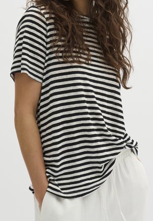 Vrouw draagt een zwart-wit gestreept T-shirt met korte mouwen en een witte broek, met handen in de zakken tegen een effen achtergrond.