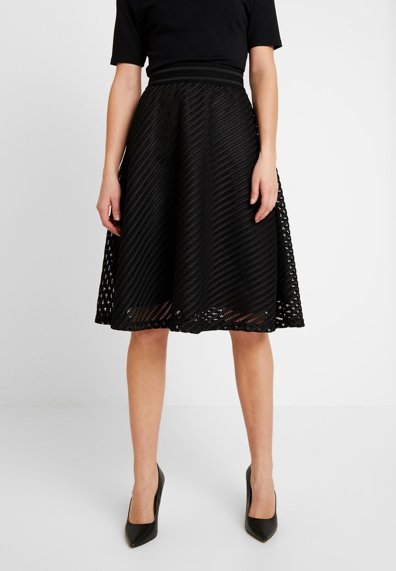 Femme portant une jupe noire texturée en forme A, un top noir à manches courtes et des talons hauts pointus noirs, debout sur un sol blanc.