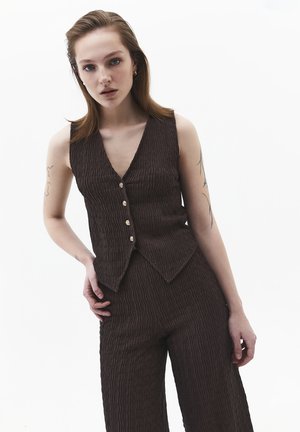 Jeune femme tatouée portant un gilet sans manches brun foncé texturé et un pantalon assorti, debout devant un fond blanc.