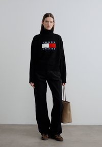 Pull noir en maille avec un écusson logo, associé à un pantalon noir. Le mannequin tient un sac à main beige et porte des baskets imprimé léopard.