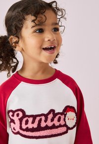Biała koszula z długim rękawem, z czerwonymi rękawami raglanowymi, z teksturowanym różowym i burgundowym napisem "Santa" oraz twarzą Świętego Mikołaja na klatce piersiowej.