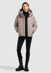 Hellbeige Steppjacke mit Kapuze, einem gesteppte Design, vorderen Knöpfen und seitlichen Taschen, kombiniert mit schwarzen Leggings und Stiefeln.