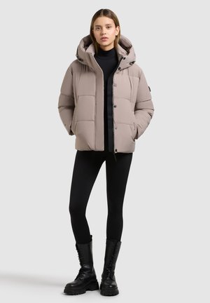 Hellbeige Steppjacke mit Kapuze, einem gesteppte Design, vorderen Knöpfen und seitlichen Taschen, kombiniert mit schwarzen Leggings und Stiefeln.