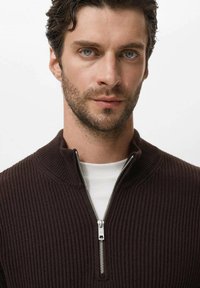 Donkerbruin geribbeld zip-up trui met een hoge kraag en zilveren rits, gelaagd over een wit shirt. Close-up weergave die de textuur van de stof benadrukt.