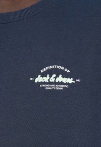 Σκούρο μπλε ύφασμα με λευκό και ανοιχτό πράσινο κείμενο που γράφει "Ορισμός Jack & Jones, Est. 1990, Ισχυρό και Αυθεντικό Ποιοτικό Denim"