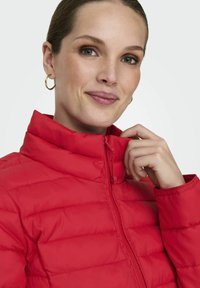 Raudonas pūkinis švarkas su aukštu apykakle, horizontaliu siuvinėjimo dizainu, užtrauktuku ir lygiu paviršiumi. Sukurtas pabrėžti figūrą.