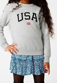 Šedá mikina s nápisem "USA" v černém písmu a malou vlajkovou emblémem, spárovaná s modrou sukní s květinovým potiskem a volány a černými punčochami.