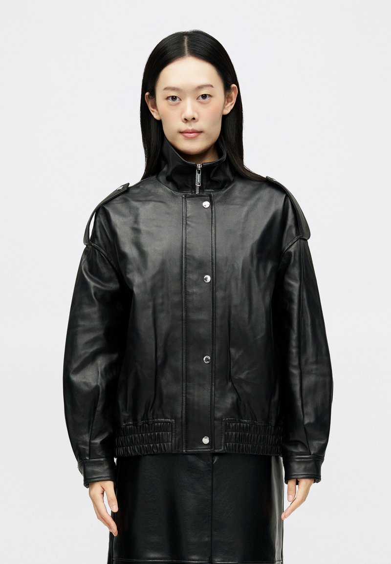 Giacca in pelle nera con colletto alto, bottoni a pressione e vita elasticizzata. Presenta dettagli sulle spalle imbottiti e maniche lunghe.