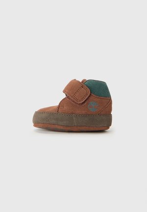 Brown suede ankle boot with a green accent, featuring a Velcro closure and a textured rubber sole for grip. Tree logo on the side.