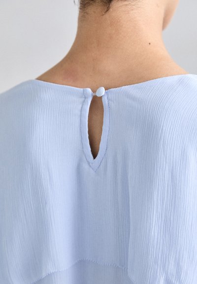 Lichtblauwe blouse met een sleutelgatdetail aan de achterkant, gestructureerde stof met subtiele plooien en een knoopsluiting.
