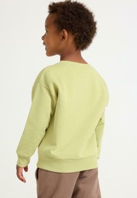 Bambino con capelli ricci che guarda di lato, indossa una felpa verde chiaro e pantaloni marroni su uno sfondo semplice.