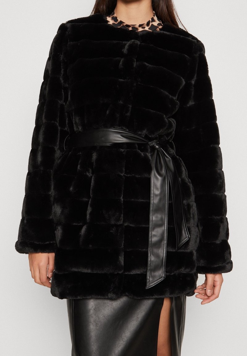 Femme portant une veste en fausse fourrure noire avec un motif matelassé, attachée avec une ceinture en cuir noire sur une jupe en cuir noire avec une fente devant.
