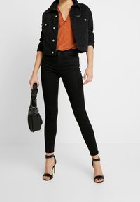 Chaqueta de mezclilla negra y vaqueros pitillo negros de talle alto, combinados con una blusa naranja y sandalias de tacón negro con tiras. Bolso cruzado negro.