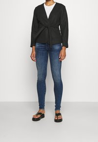 Chaqueta envolvente negra de manga larga con cinturón en la cintura, combinada con jeans ajustados azules y sandalias negras tipo deslizante sobre un fondo liso.