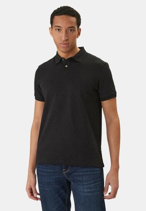 Giovane uomo in piedi, indossa una polo nera a maniche corte e jeans blu, su uno sfondo bianco semplice.