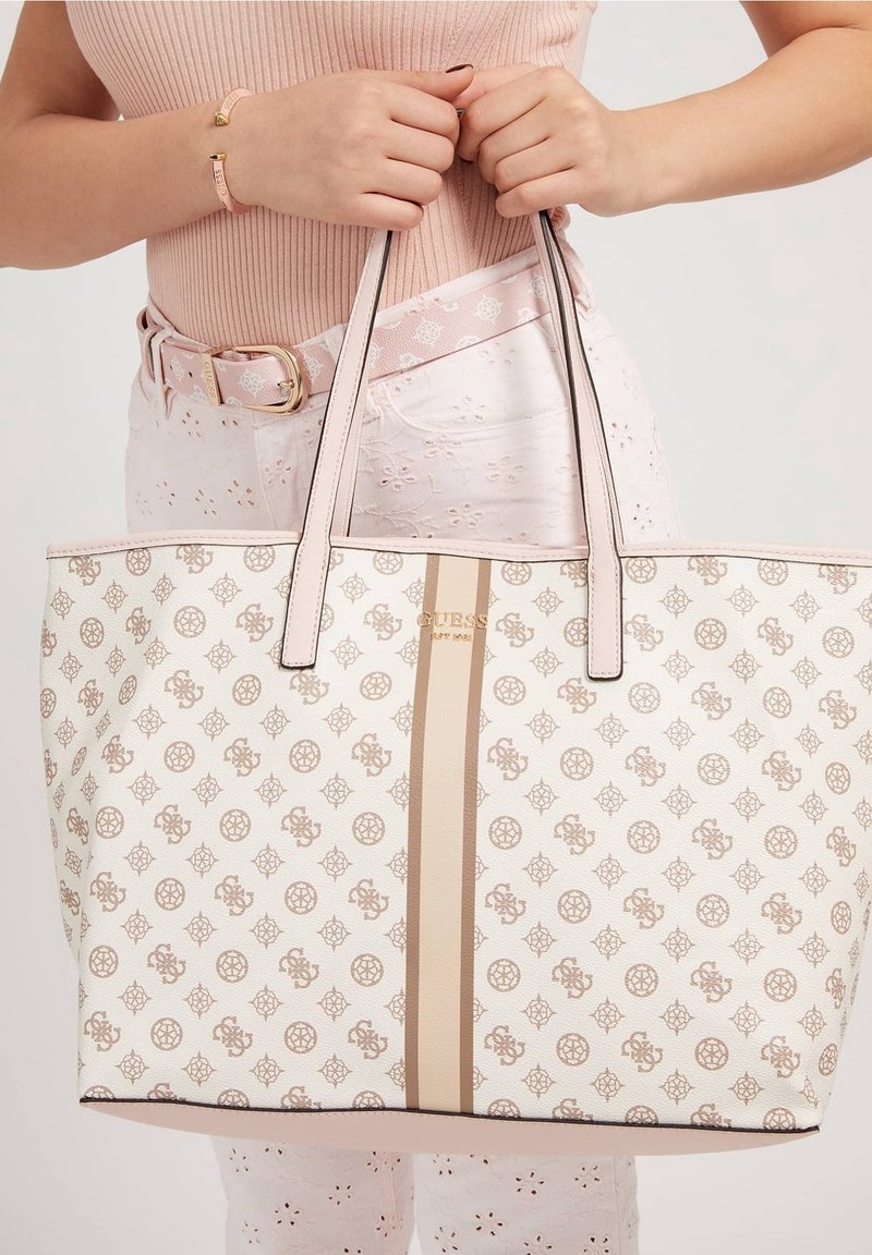 Femme tenant un sac fourre-tout beige à motifs avec deux bandes verticales, portant un haut côtelé rose, un pantalon à œillets, une ceinture et un bracelet assortis.