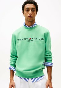 Sweat-shirt vert avec le texte "TOMMY HILFIGER EST. 1985", présentant un col rond et des poignets côtelés, superposé sur une chemise à rayures bleues et blanches.
