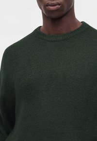Gros plan d'une personne portant un pull en maille texturé vert foncé à col rond sur un fond clair uni.