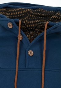 Blouson à capuche bleu avec cordon de serrage marron, boutons en bois et doublure intérieure à motifs dans des tons foncés et clairs. Fermeture éclair à l'avant.