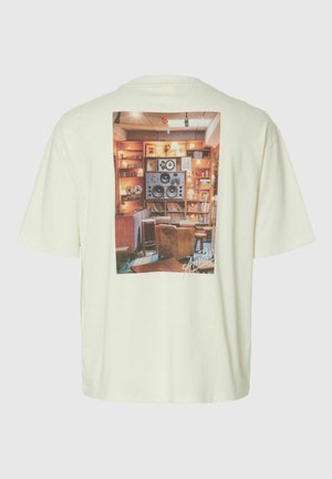 Krem T-skjorte med et trykt bilde av et koselig rom med store høyttalere, bokhyller, stoler og varm belysning.