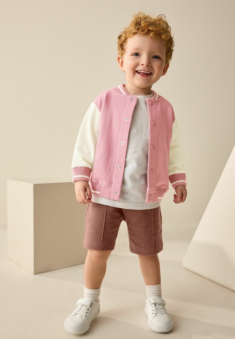 Unge barn med krøllet rødt hår, smilende, iført en pink og hvid jakke, hvid skjorte, brune shorts, hvide sokker og hvide sneakers.