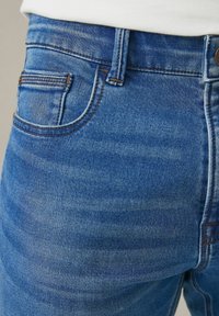 Acercamiento a la zona de la cintura de unos jeans de mezclilla azules, mostrando el bolsillo frontal, el lazo del cinturón, el botón y la textura de la tela con franjas horizontales desgastadas.