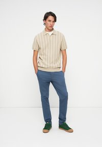 Beige striped polo shirt with short sleeves, paired with blue trousers and green sneakers with brown soles. Model standing, hands in pockets.
