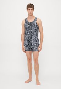 Junger Mann in einem passenden Outfit aus schwarz-grauem Leopardenmuster: Tanktop und Shorts, steht barfuß vor einem schlichten weißen Hintergrund.