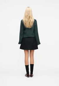 Femme blonde debout, de dos, portant un haut à manches longues vert foncé, une jupe noire, des chaussettes noires et des chaussures marron sur fond blanc.