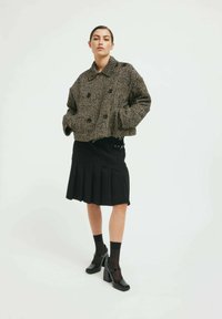 Jachetă croită cu strat dublu, în culoare maro și negru, cu model herringbone, asociată cu o fustă plisată neagră și pantofi cu toc gros negri. Material texturat.