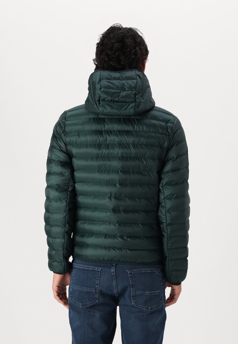 Chaqueta de plumas verde oscuro con diseño acolchado horizontal, capucha y tejido suave. Llevada sobre jeans azules, mostrando la vista trasera.