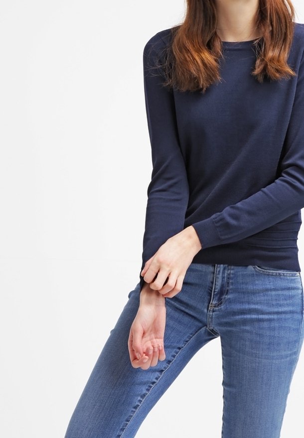 Marineblaues Strickpullover mit langen Ärmeln und rundem Halsausschnitt, ergänzt durch eine taillierte Passform und eine glatte Textur. Kombiniert mit blauen Jeans.