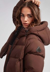 Brązowa kurtka puchowa z dużym kapturem, z czarnym zamkiem błyskawicznym oraz trójkątną łatą z logo na lewym rękawie. Gładka, pikowana tekstura.