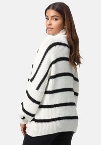 Pull en maille épaisse blanc avec des rayures noires, doté d'un col montant, de manches larges et d'une texture côtelée. Coupe ample, style décontracté.