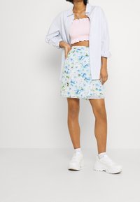 Himmelblaues Hemd in Button-Up-Style über einem pinken, gerafften Crop-Top, kombiniert mit einem Rock mit floralen Mustern in Blau- und Grüntönen sowie weißen Sneakers.