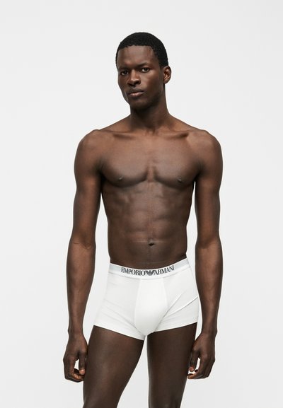 Άνδρας μοντέλο με σκουρόχρωμο δέρμα, που φοράει λευκά boxer briefs Emporio Armani, στέκεται μπροστά σε ένα μονόχρωμο ανοιχτό γκρι φόντο.