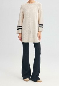 Abito maglione lavorato a maglia crema con motivo geometrico e accenti a strisce nere sulle maniche, abbinato a pantaloni neri aderenti e scarpe beige.