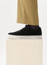 Sorte mockasinsneaker med hvit gummisåle, med rund tå, snøring og strukturert overflate, plassert på en lys blokk.