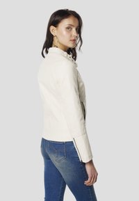 Biela kožená bunda so priliehavým strihom, stojacím golierom, zipsovými manžetami a bočnými vreckami, v kombinácii s modrými džínsami z denimu.