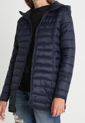 Femme portant une veste matelassée à capuche bleu marine sur un top noir et un jean bleu déchiré, debout devant un fond blanc.