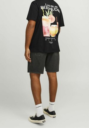 Person iført sort Bermuda-grafik T-shirt, sorte denimshorts, hvide sokker og sorte lavtop-sneakers, stående mod en ensfarvet baggrund.
